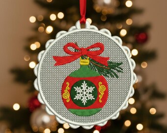 May include: Embroidered Christmas ornament with a red bow and a green and red bauble design. The ornament features a snowflake and decorative gold swirls. It hangs from a red ribbon against a blurred Christmas tree background.