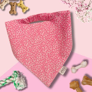 Könnte beinhalten: Rosa Hunde-Bandana mit Blumenmuster und elastischem Nackenband. Das Bandana besteht aus hellrosa Stoff mit einem Muster aus kleinen, dunkelrosa Blüten. Hundeleckerlis und ein Seilspielzeug sind ebenfalls zu sehen.