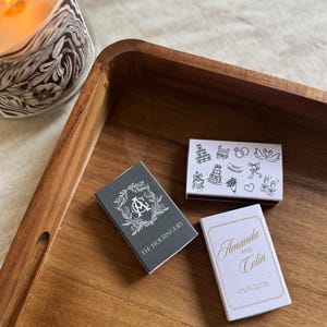 May include: Three matchboxes on a wooden tray. One grey matchbox has a monogram and the text "THE HOLSINGERS". A white matchbox has the names "Amanda and Colin" and the date "05.16.2026". The third matchbox has wedding-themed illustrations.
