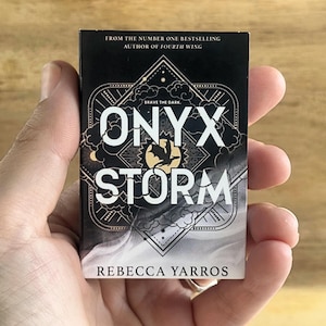 May include: A small book titled "ONYX STORM" by Rebecca Yarros is held in a hand. The cover is black with white and gold accents, featuring the title in large letters and a graphic design. The text "FROM THE NUMBER ONE BESTSELLING AUTHOR OF FOURTH WING" is at the top.