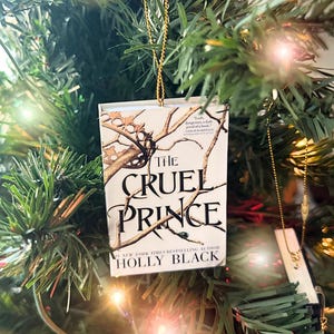 May include: A Christmas tree ornament shaped like a book cover, featuring the title "The Cruel Prince" by Holly Black. The cover art shows a branch with a crown and the book is suspended by a gold string.