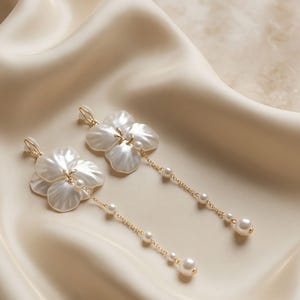 Könnte beinhalten: Ein Paar goldfarbene Ohrhänger mit weißen, blütenblattförmigen Elementen. Jeder Ohrring besteht aus einer zarten Goldkette, die von der Blume herabhängt und mit kleinen, runden weißen Perlen besetzt ist, die entlang der Kette verteilt sind.