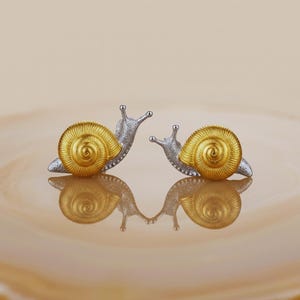 Könnte beinhalten: Ein Paar schneckenförmige Ohrstecker mit einem texturierten silbernen Körper und einem leuchtend goldenen Spiralgehäuse. Die beiden Schnecken sind einander zugewandt und zeigen eine detaillierte Metallverarbeitung mit poliertem Finish.