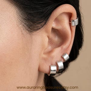 Könnte beinhalten: Ein Set aus vier polierten silberfarbenen Ohrklemmen und Creolen, die am Ohr getragen werden. Der Schmuck zeichnet sich durch ein glattes, breites und geschwungenes Banddesign aus. Der Text www.auraringtreasures.etsy.com steht unten im Bild.