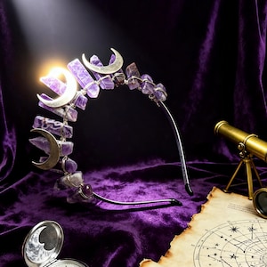 May include: A crown with amethyst crystals and silver crescent moon details. The headpiece sits on a thin silver band. The image is set against a purple velvet backdrop, with a pocket watch, telescope, and a star chart.