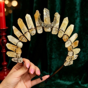 May include: A crystal crown with clear and yellow quartz crystals. The crystals are arranged in an arc, held together with wire and a decorative crescent moon. The crown is held by a hand with pink nail polish.