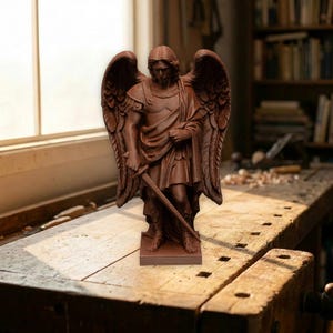 Puede incluir: Una estatua marrón de un ángel con grandes alas, sosteniendo una espada. La estatua está sobre una base rectangular. El ángel lleva una armadura y una túnica fluida. El fondo incluye un banco de trabajo de madera y una estantería.