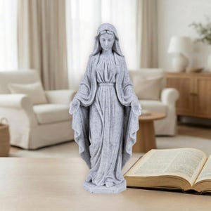 Może przedstawiać: Szara statua kobiety z wyciągniętymi ramionami, ubranej w drapowaną szatę i nakrycie głowy. Statua ma nakrapianą fakturę i stoi na okrągłej podstawie. Statua stoi na drewnianym stole.