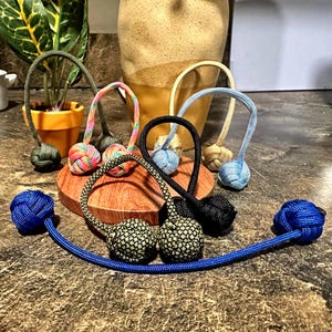 May include: A collection of colorful monkey fist keychains. The keychains feature various colors, including blue, black, green, and rainbow patterns. Each keychain has a knotted ball design and a loop for attachment.