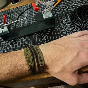 Puede incluir: Dos pulseras verde oliva y negras se llevan en la muñeca. Las bandas verde oliva tienen la palabra "SNIPER" impresa. Las pulseras están en el brazo de una persona, con una esterilla de corte negra y herramientas en el fondo.