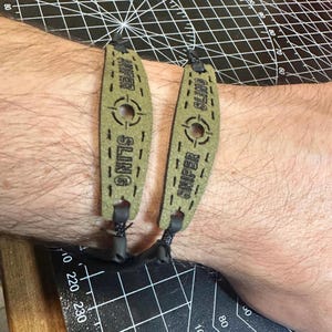 May include: Two olive green slingshot wrist braces with the text "SNIPER SLING" and a target symbol. The braces are connected with black cords. The background is a black cutting mat.