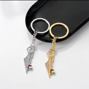 May include: Two keychains, one silver and one gold, each featuring a map-shaped pendant with sparkling embellishments. The pendants include a small flag design. The keychains are displayed on a black surface.