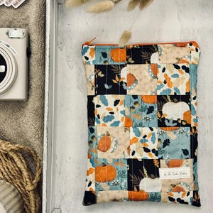 Peut inclure: Pochette matelassée sur le thème de l'automne avec un motif patchwork représentant des citrouilles, des feuilles et des motifs floraux orange, bleus et crème. La pochette a une fermeture éclair orange et une étiquette "Handmade by The Kindred Studio."