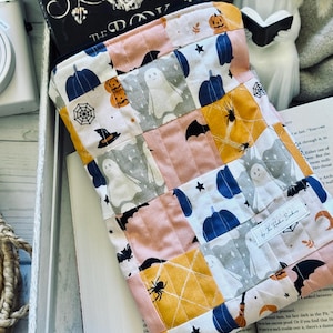 May include: A Halloween-themed book sleeve with a patchwork design. The sleeve displays ghosts, pumpkins, bats, and spiders in pink, grey, yellow, and blue. A book with a skull on the cover is partially visible, along with the text "THE DO".