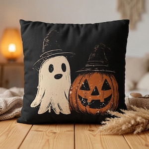 May include: A square black throw pillow featuring a graphic illustration of a white ghost and an orange jack-o'-lantern, both wearing black pointed witch hats. The design has a distressed, textured appearance with clean lines and high contrast.