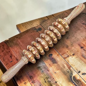 Puede incluir: Un rodillo de masaje de madera con una serie de cilindros rodantes. Los cilindros y los mangos son de madera clara con patrones de vetas rojas y marrones. La herramienta está diseñada para la terapia muscular y la relajación.