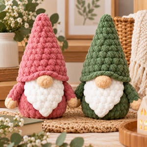 May include: Two plush gnome figurines crafted from chunky yarn. One gnome features a dusty rose pink hat and body, while the other has a forest green hat and body. Both gnomes have a round beige nose and a textured white beard made of soft yarn.