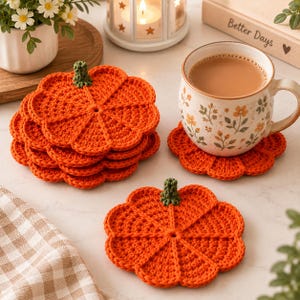Puede incluir: Un juego de posavasos de ganchillo con forma de calabaza naranja y tallos verdes. Un posavasos está plano, mientras que una pila de otros cuatro está cerca. Una taza de cerámica con un estampado floral descansa sobre uno de los posavasos.
