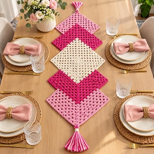 May include: A crochet table runner featuring a series of connected square motifs in shades of light pink, dark pink, and cream. The runner is arranged vertically down the center of a table and is finished with decorative tassels at both ends.