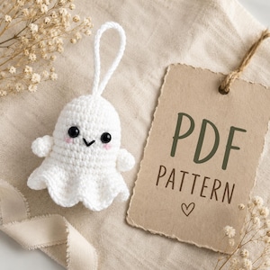 Könnte beinhalten: Ein kleines weißes gehäkeltes Geisterornament mit schwarzen Perlenaugen, einem gestickten schwarzen Mund und rosa Wangen. Ein beigefarbenes Papieretikett ist befestigt, das den dunkelgrünen Text "PDF PATTERN" und ein kleines Herzsymbol zeigt.