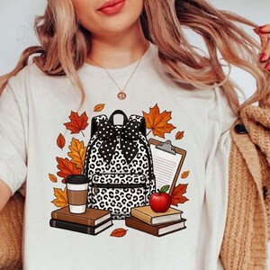 Pode incluir: Uma t-shirt cor de creme com uma impressão gráfica de uma mochila com padrão de leopardo e um laço de bolinhas preto e branco, rodeada por folhas de outono, uma prancheta, uma maçã vermelha, um copo de café e pilhas de livros em tons castanhos.