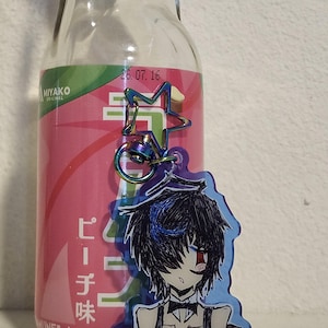 May include: A clear glass bottle with a pink label and Japanese text is next to a cartoon keychain. The keychain features a drawing of a person with black hair, a bow tie, and a bandage on their face. A star-shaped charm is attached to the keychain.