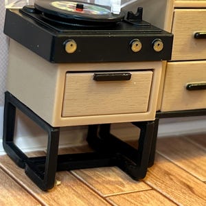 May include: Miniature record player with a black top and a beige cabinet. The record player has a clear plastic cover and a colorful record on the turntable. The cabinet has a drawer with a black handle and sits on a black metal frame.