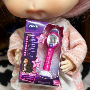May include: A pink and white VTech Kidi Star Music Magic Microphone toy in its original packaging. The box features a photo of a girl holding the microphone and lists features like built-in songs and voice-changing effects. The toy is designed for children aged 5 and up.