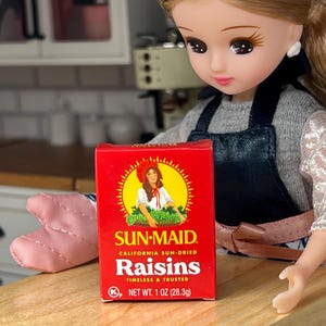 May include: A miniature box of Sun-Maid California Sun-Dried Raisins. The red box features a woman in a bonnet and the text "Raisins". The net weight is 1 oz (28.3g).