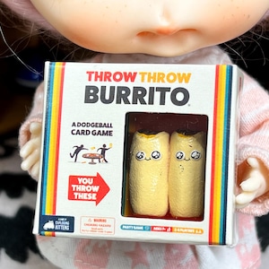 May include: A small box of the card game "Throw Throw Burrito" is held up. The box has a colorful rainbow border and features two cartoon burritos with smiling faces. Text on the box reads "A Dodgeball Card Game".