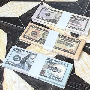 Może przedstawiać: Trzy stosy banknotów dolarów amerykańskich, każdy owinięty białą papierową opaską. Nominały to 50 $, 10 $ i 100 $. Banknoty leżą na wzorzystej powierzchni z czarnymi, beżowymi i brązowymi kształtami geometrycznymi.