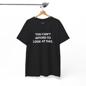 May include: Black t-shirt with the white text "YOU CAN'T AFFORD TO LOOK AT THIS." The shirt is hanging on a wooden hanger attached to a metal rod. The shirt is a basic crew neck style.