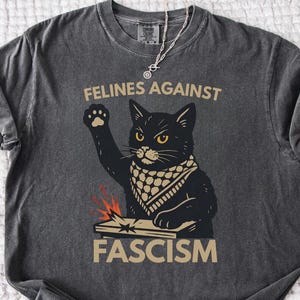May include: Pepper-colored t-shirt featuring a black cat illustration with a raised paw, wearing a bandana, and the text "FELINES AGAINST FASCISM." The design includes a stylized image of a book and sparks. The shirt is unisex.