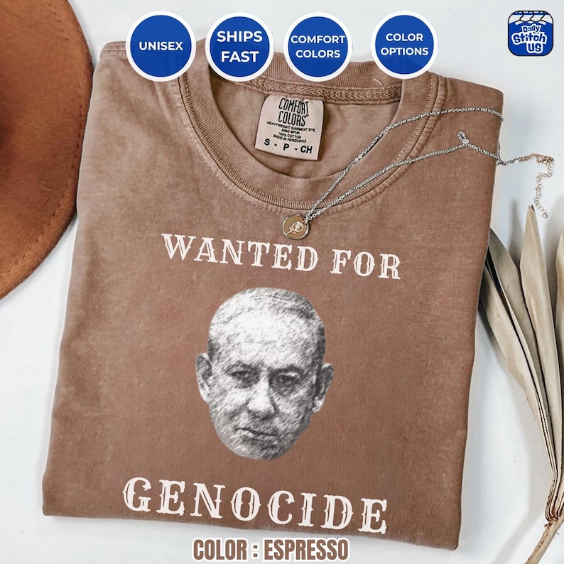 May include: Espresso-colored unisex t-shirt with the text "WANTED FOR GENOCIDE" and a grayscale portrait. The shirt features a crew neck and is made of Comfort Colors material. A silver necklace with a pendant is draped across the shirt.