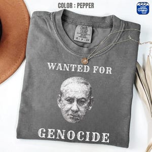 May include: Pepper-colored t-shirt with the text "WANTED FOR GENOCIDE" above and below a grayscale portrait. The shirt has a crew neck and a small tag. A gold necklace with a pendant is draped across the shirt.