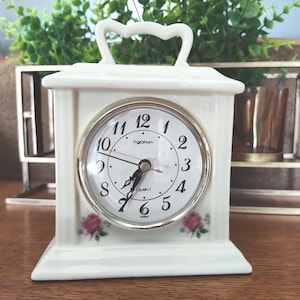 May include: A cream-colored quartz clock with a silver-tone bezel and a white face. The clock has black numerals and hands, with small pink rose accents. The clock has a handle and is sitting on a wooden surface.