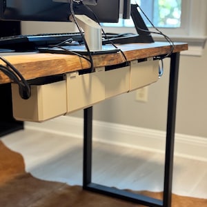 May include: A wooden desk with a black metal frame. Beige cable management trays are attached underneath the desk. A computer monitor and keyboard are on top. The desk sits on a brown and white rug.
