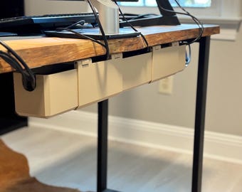 May include: A wooden desk with a black metal frame. Beige cable management trays are attached underneath the desk. A computer monitor and keyboard are on top of the desk. The desk sits on a brown and white rug.