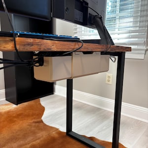 May include: A computer desk with a dark wood top and black metal legs. A beige cable management tray is mounted under the desk. A black monitor and keyboard are visible. The desk sits on a brown cowhide rug.