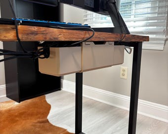 May include: A computer desk with a dark wood top and black metal legs. A beige cable management tray is mounted under the desk. A black monitor and keyboard are visible. The desk sits on a brown cowhide rug.