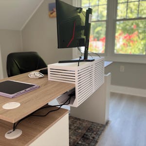 Puede incluir: Una caja blanca de gestión de cables con listones se encuentra sobre un escritorio de madera clara. La caja oculta cables y regletas, con un diseño de rejilla horizontal. Un monitor de computadora negro se apoya encima, junto a una tableta y un ratón.