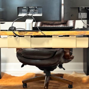 May include: A cream-coloured under-desk cable tidy is mounted beneath a wooden desk. The desk supports two computer monitors, with cables neatly organised. A brown leather office chair sits below the desk, and a cowhide rug is on the floor.
