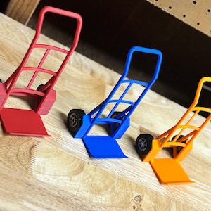 Może przedstawiać: Trzy miniaturowe wózki ręczne w kolorze czerwonym, niebieskim i pomarańczowym ułożone na drewnianej powierzchni. Każdy wózek ma prostokątną podstawę, pionową ramę i zakrzywiony uchwyt. Koła są czarne.