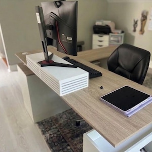 May include: A light oak L-shaped desk featuring a white monitor stand with horizontal slats, a black computer monitor, a black keyboard, a black office chair, and a tablet in a light purple case resting on the desk surface.