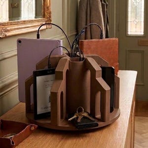 May include: A brown, circular, multi-slot wooden charging station with a central hub for cables. It holds a light purple tablet, a brown leather-covered device, and a black e-reader. A set of metal keys rests in one of the open storage sections.