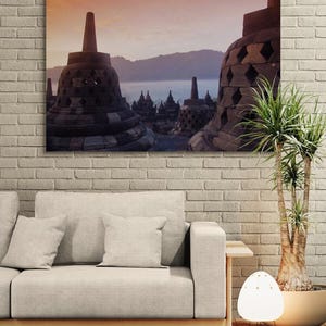 Op de afbeelding: Een ingelijste canvas print van het Borobudur tempelcomplex in Indonesië. De afbeelding toont de oude boeddhistische tempel met zijn iconische stoepa's en ingewikkeld metselwerk, tegen een achtergrond van bergen en een schemerige lucht. De print wordt boven een lichtgekleurde bank weergegeven.