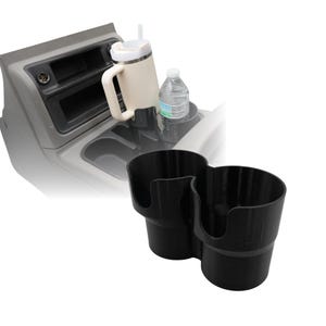 Puede incluir: Un portavasos doble negro diseñado para la consola central de un vehículo. El soporte está hecho de un material negro mate sólido y presenta dos compartimentos circulares con un divisor de conexión. Una taza beige con asa y una botella de agua están en el fondo.