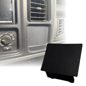 May include: A black, textured plastic panel, likely an automotive interior component, is shown. The panel is rectangular with a slightly angled top surface. The background shows a vehicle's dashboard with air vents and control knobs.
