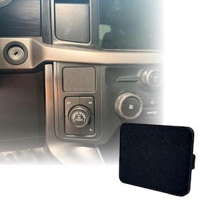May include: A rectangular black plastic cover with a textured surface designed to fit into the dashboard slot of a vehicle. The cover is shown next to a vehicle interior console featuring a drive mode dial, climate control knobs, and buttons.
