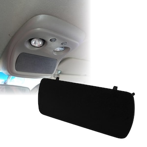 Puede incluir: Una cubierta negra para la consola superior de un vehículo. La cubierta es rectangular con bordes redondeados y tiene dos pequeñas pestañas en el borde superior. El fondo muestra el interior de un vehículo, incluyendo luces y un compartimento de almacenamiento.